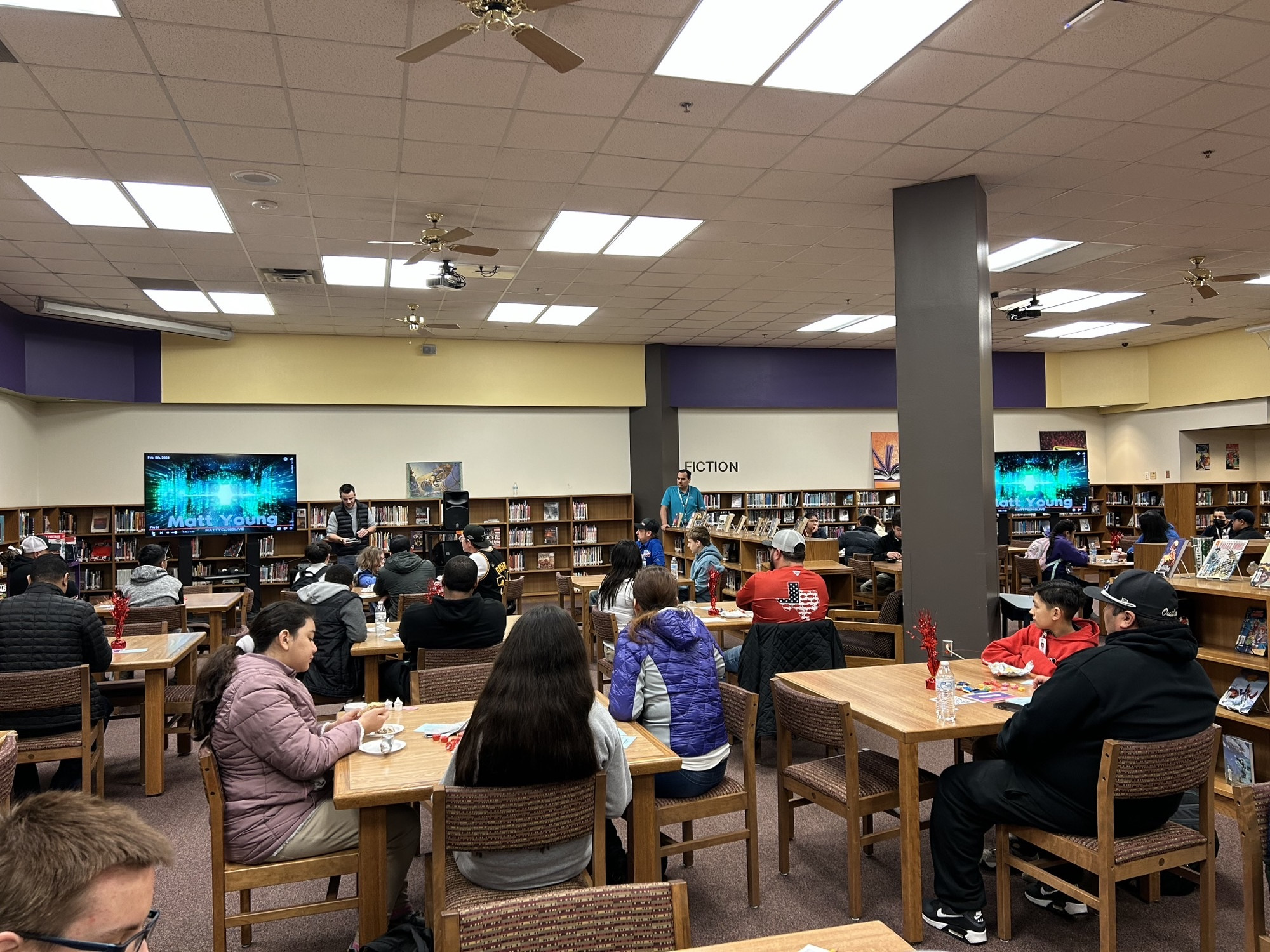 Matt Young speaking to a packed school library of dads and kids at an All Pro Dad event, his name on the screens