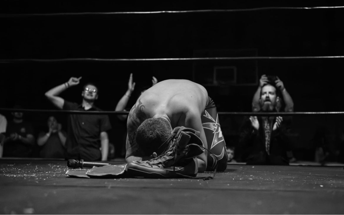 Black-and-white photo of Matt Young kneeling over his boots in the ring after his final match
